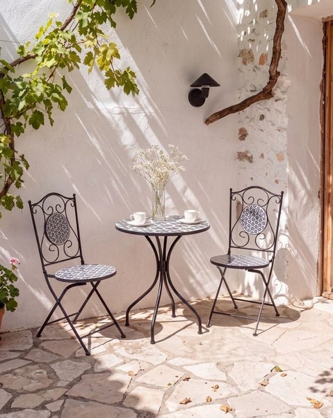 Mosaik Bistroset bestehend aus einem runden Tisch und zwei Klappstühlen mit dekorativen Mustern auf einer Terrasse vor einer weißen Mauer.