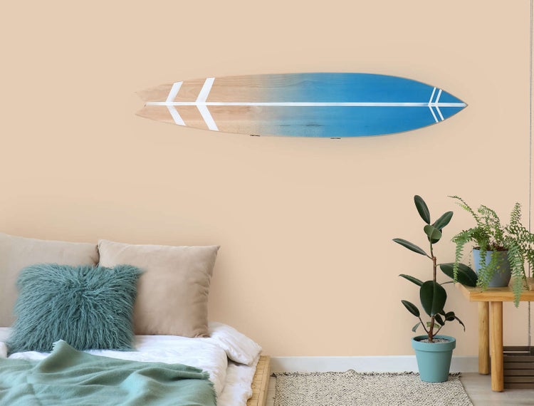 Wanddekoration in Form eines Surfbretts aus Holz über einem Bett in einem hellen Schlafzimmer mit Zimmerpflanzen.