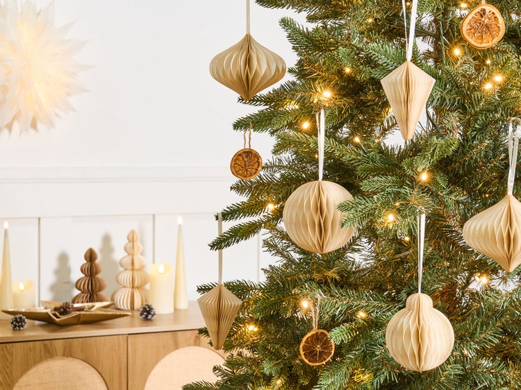 Geschmückter Weihnachtsbaum mit Papieranhängern in Wabenoptik, Orangen und Lichterkette in einem hellen Raum mit Holzmöbeln und Kerzen.