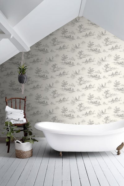 Badezimmer mit Toile-de-Jouy-Tapete, freistehender Badewanne, Holzstuhl mit Handtüchern und Pflanzen auf Holzdielen.