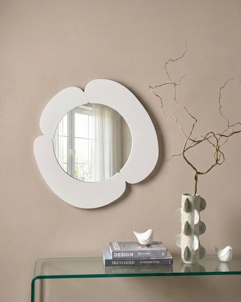 Hell gestalteter Raum mit einem weißen, blumenförmigen Wandspiegel über einer Glaskonsole mit Dekorationselementen wie Büchern und einer Vase.