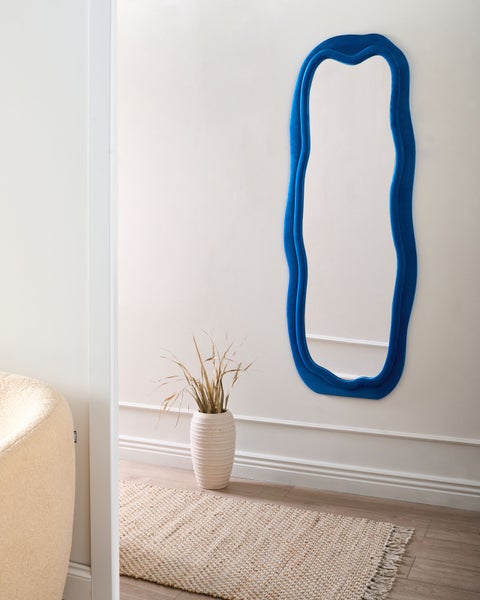 Moderner blauer Wandspiegel mit gewelltem Rahmen in einem hellen Raum mit Vase und Teppich.