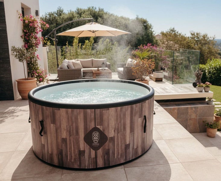 Aufblasbarer Whirlpool mit COCO SPA Logo in Holzoptik und sprudelndem Wasser auf einer hellen Terrasse, im Hintergrund Loungebereich mit Sonnenschirm.