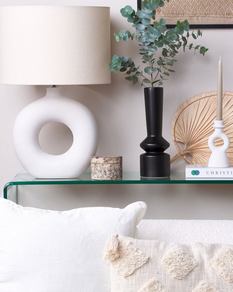 Dekoratives Interieur mit Lampe, Vase, Kerzenhalter und Kissen auf einem Glastisch.