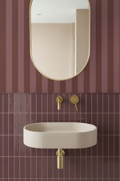 Modernes Badezimmer mit beigem ovalem Aufsatzwaschbecken, goldener Wandarmatur und Kapselspiegel vor bordeauxroten Fliesen und Streifentapete.