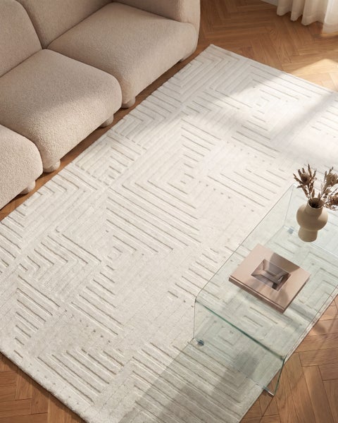 Wohnzimmer mit strukturiertem Webteppich mit geometrischem Muster, davor ein Sofa und ein Glastisch auf Parkettboden.