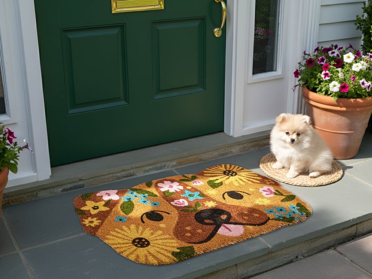 Farbenfrohe Fußmatte in Form eines Hundekopfs mit Blumenmuster vor einer grünen Haustür auf einer Steinterrasse mit kleinem Hund.