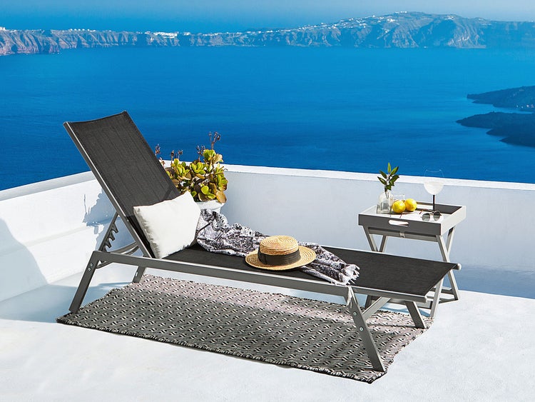 Gartenlounge mit Liegestuhl, Beistelltisch und Teppich vor einer Meereskulisse.