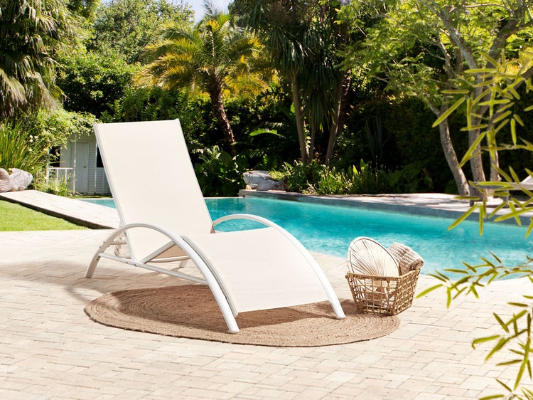 Moderne Sonnenliege mit geschwungenem Rahmen auf einem runden Teppich am Pool, umgeben von einem hellen tropischen Garten.
