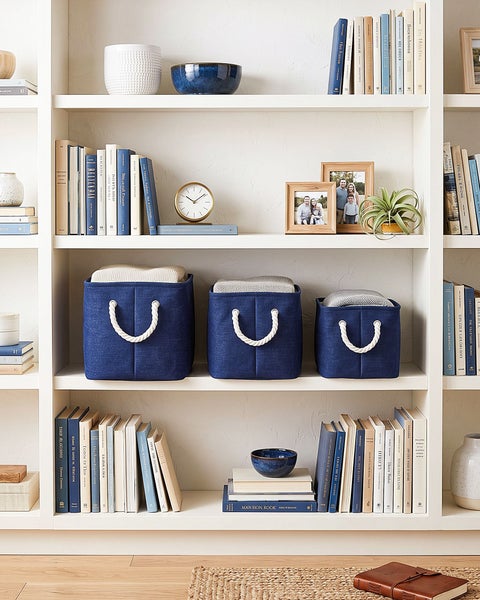 Drei textile Aufbewahrungskörbe in verschiedenen Größen mit Seilgriffen in einem weißen Regal, dekoriert mit Büchern und Wohnaccessoires.