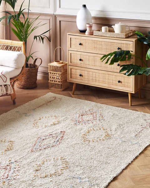 Hochflor-Teppich mit Rautenmuster in einem hellen Wohnzimmer mit Kommode aus Holz und Rattan, einem Rattan-Sessel und Zimmerpflanzen.