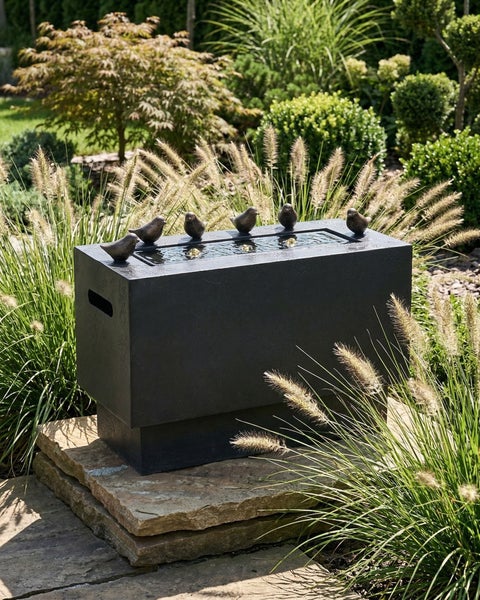 Rechteckiger schwarzer Gartenbrunnen mit fünf Vogelfiguren am Rand, platziert auf Natursteinplatten inmitten von Ziergräsern in einem Garten.