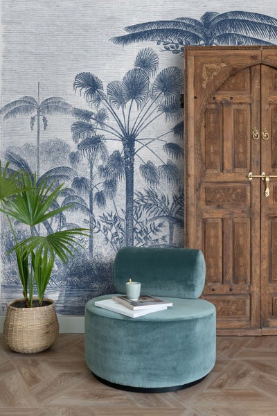 Vliestapete mit blauem Palmenmuster im Vintage-Stil in einem Wohnraum mit Samtsessel und antikem Holzschrank.