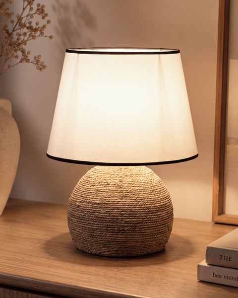 Tischlampe mit rundem Fuß aus geflochtenem Seil und klassischem Lampenschirm auf einem Holztisch als stimmungsvolle Wohnzimmerbeleuchtung.