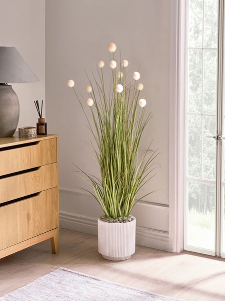 Dekorative Kunstpflanze im Topf neben Kommode und Fenster.