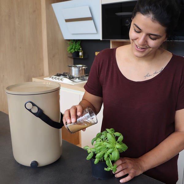 Frau gießt Flüssigkeit aus einem Glas in einen Blumentopf neben einem Komposteimer in der Küche