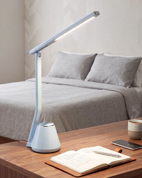 Moderne LED Schreibtischleuchte mit verstellbarem Leuchtenarm auf einem Holztisch vor einem Bett im Schlafzimmer.