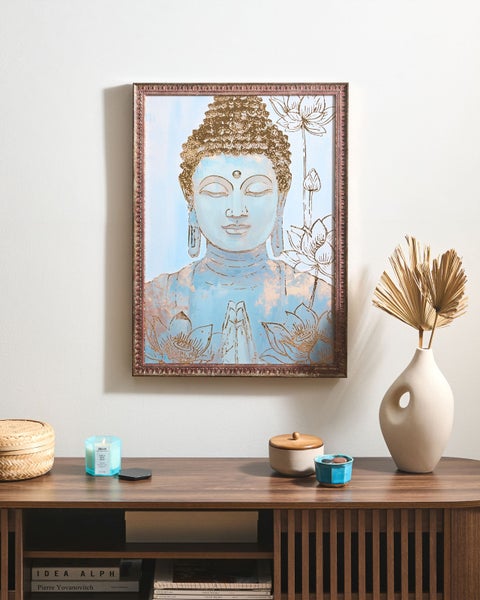 Gerahmtes Wandbild mit Buddha-Motiv und Lotosblüten über einem Sideboard mit dekorativen Wohnaccessoires wie Vase und Kerze.