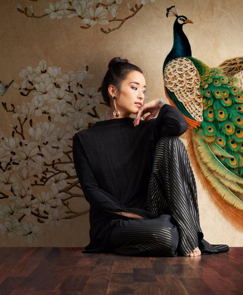 Frau sitzt vor einer Tapete mit Pfauen- und Blumenmuster.
