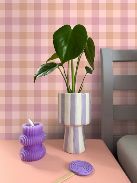 Grüne Monstera in einer lila-weiß gestreiften Keramikvase neben einer lila Kerze auf einem rosa Tisch vor einer rosa-weiß karierten Tapete.