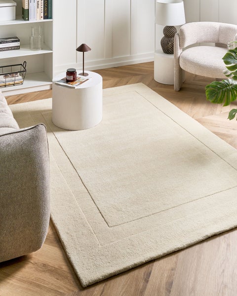 Moderner cremefarbener Teppich mit Relief-Bordüre in einem hellen Wohnzimmer auf Parkettboden neben einem Sofa und einem Sessel.