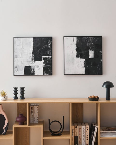 Zwei quadratische abstrakte Wandbilder in Schwarz-Weiß mit schwarzem Rahmen in einem modern eingerichteten Raum über einem Holzregal.