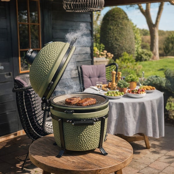 Grüner Keramikgrill Kamado auf einem runden Holztisch im Garten mit geöffnetem Deckel und grillendem Fleisch.