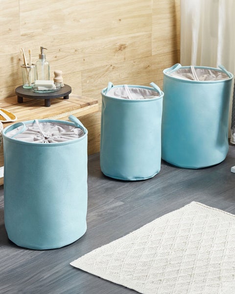 Drei hellblaue Wäschekörbe aus Stoff mit Kordelzug in verschiedenen Größen in einem hellen Badezimmer mit Holzdekor und weißem Teppich.