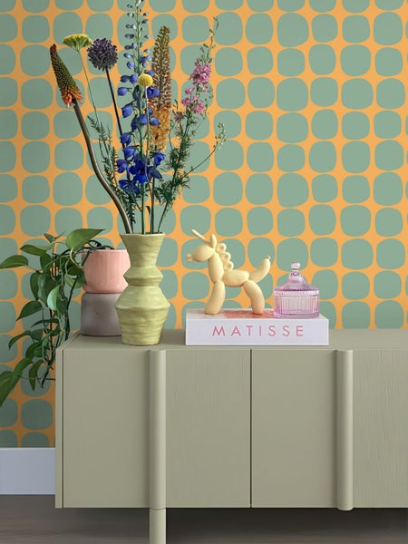 Vliestapete mit retro-geometrischem Muster aus abgerundeten Quadraten an einer Wand hinter einem Sideboard mit Dekoration und Blumen.