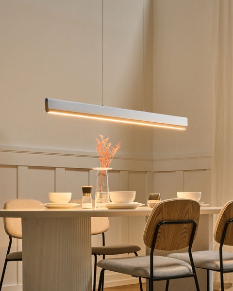 Lange lineare LED-Pendelleuchte über einem weißen Esstisch mit Holzstühlen in einem hellen, modern gestalteten Raum.