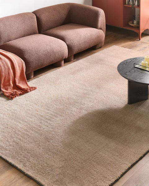 Hochfloriger beiger Teppich in einem modernen Wohnzimmer mit braunem Bouclé-Sofa, dunklem Couchtisch und Holzfußboden.