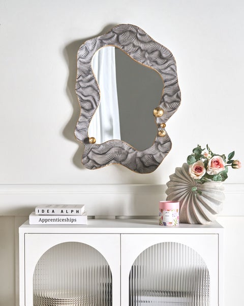 Dekoratives Interieur mit einem Spiegel mit unregelmäßigem Rahmen, einer Blumenvase und dekorativen Büchern auf einem weißen Schrank.