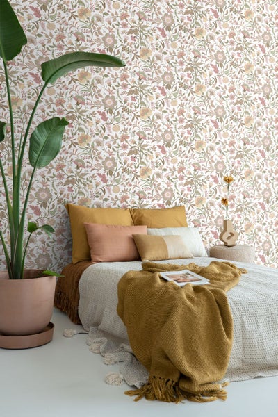 Helles Schlafzimmer mit Blumentapete, Bett mit Kissen in warmen Tönen, strukturierter Tagesdecke und einer großen Pflanze im Tontopf.