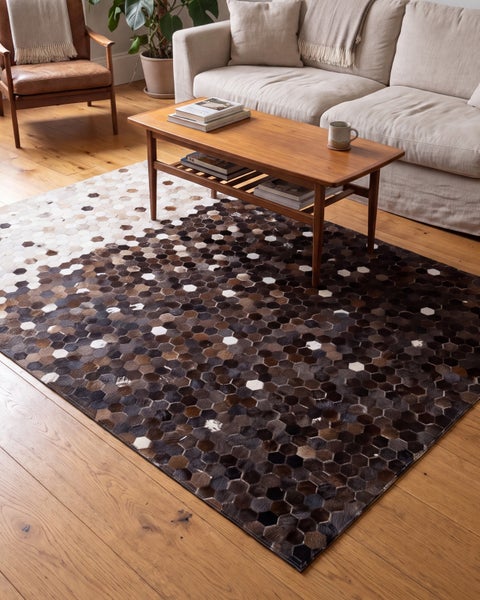 Wohnzimmer mit beigem Sofa und Couchtisch auf einem rechteckigen Teppich aus Fellstücken in Sechseckform mit Farbverlauf auf Holzboden.