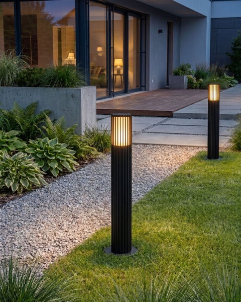 Moderne schwarze Pollerleuchten mit warmweißem Licht an einem Gartenweg aus Kies neben einer Rasenfläche am Abend.