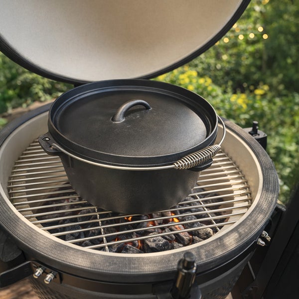 Gusseiserner Dutch Oven mit Deckel auf einem Edelstahl-Grillrost in einem Keramikgrill über glühenden Kohlen.