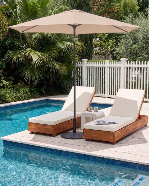 Heller Gartenbereich mit Sonnenschirm und zwei Sonnenliegen aus Holz am Pool sowie Zaun und tropischen Pflanzen im Hintergrund.