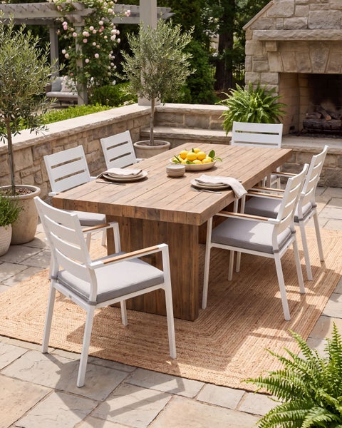 Gartenessgruppe mit einem Holztisch und sechs Stühlen mit Sitzpolstern auf einer Terrasse vor einem Steinkamin im Garten.