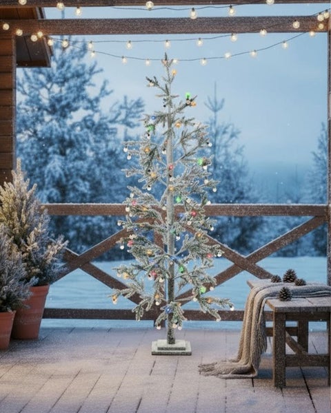 Ein künstlicher, beschneiter Weihnachtsbaum mit Lichterkette auf einer winterlichen Terrasse im Freien.