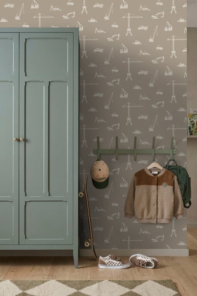 Kinderzimmer mit Baustellentapete, grünem Kleiderschrank, Garderobe mit Kleidung und einem Skateboard vor einer Wand mit Kranmotiven.