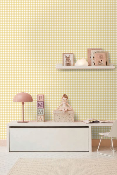 Helles Kinderzimmer mit gelb-weiß karierter Tapete, weißem Schreibtisch mit rosa Lampe, Buchstabenwürfeln, Puppe und einem Wandregal mit Büchern.