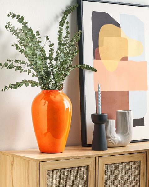 Dekorative Szene mit orangefarbener Vase, Pflanzen, Kerzenständer und abstraktem Bild auf einem Holzschrank.