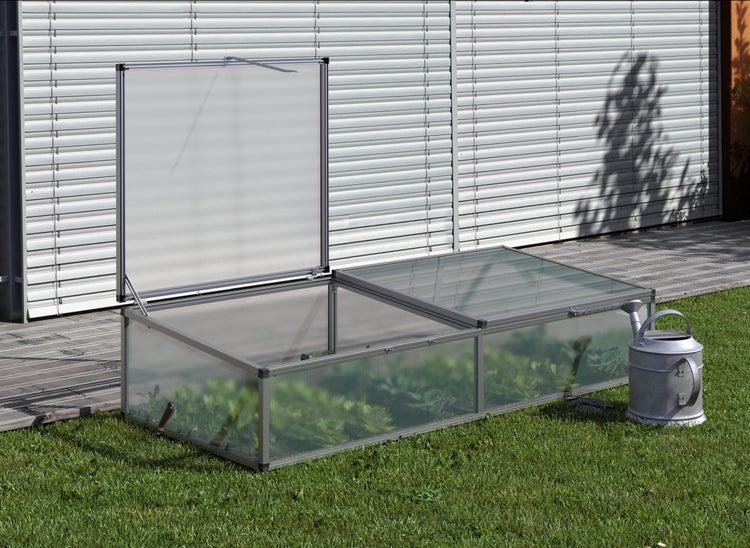 Frühbeet aus Aluminium mit Hohlkammerplatten, zwei Klappdeckeln, einer geöffnet, auf einer Rasenfläche neben einer Gießkanne.