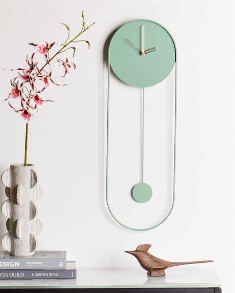 Moderne Wanduhr mit Pendel aus Metall neben einer Vase mit Orchideen und einer hölzernen Vogelfigur in einem hellen, aufgeräumten Ambiente.
