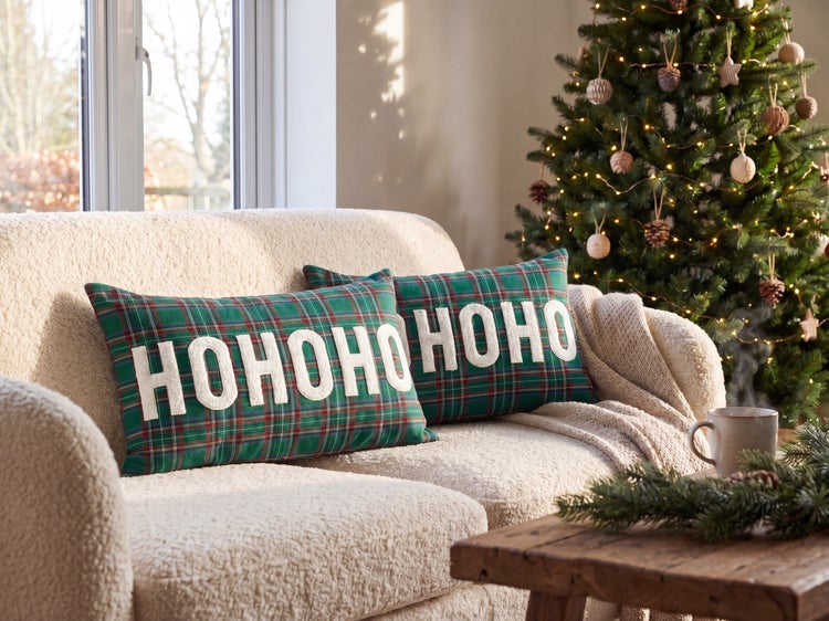 Zwei karierte Weihnachtskissen mit weißem HOHOHO Schriftzug auf einem hellen Sofa neben einem festlich geschmückten Weihnachtsbaum.