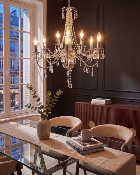 Ein eleganter Kristallkronleuchter beleuchtet ein modernes Esszimmer mit einem Glastisch und gepolsterten Stühlen vor einem Fenster.
