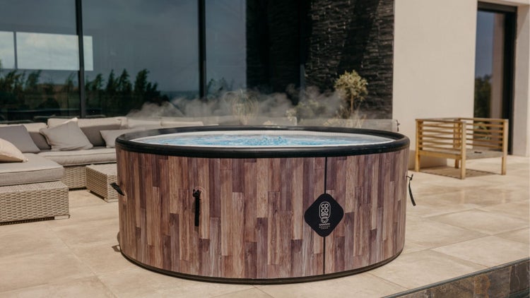 Runder Whirlpool mit Holzoptik-Verkleidung auf einer Terrasse mit Rattan-Gartenmöbeln und großen Glasfronten.