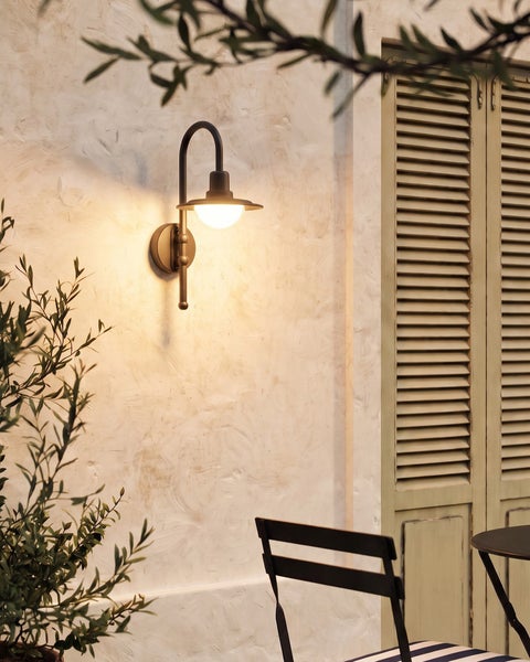 Leuchtende Außenwandleuchte im klassischen Design an einer verputzten Wand auf einer Terrasse mit Pflanze und Stuhl.
