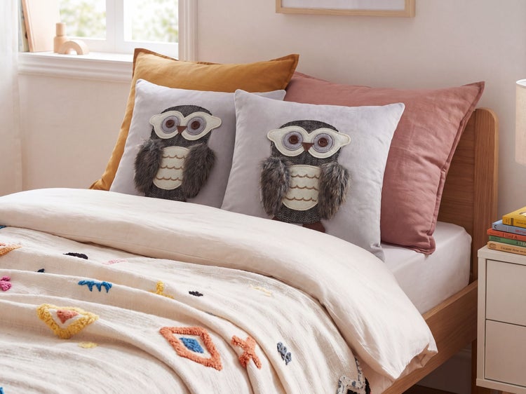 Hellgraue Dekokissen mit Eulen-Motiv und Kunstfellflügeln auf einem Holzbett in einem hellen Schlafzimmer mit gemusterter Wohndecke.