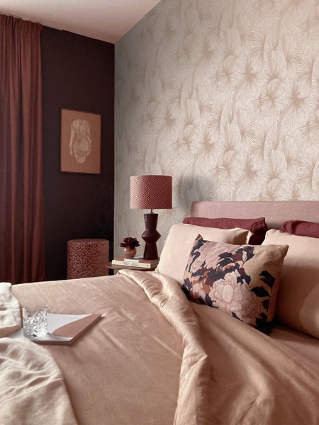 Schlafzimmer mit Mustertapete mit Blattmotiven, Bett mit Kissen in verschiedenen Mustern und einer Tischlampe auf einem Nachttisch.
